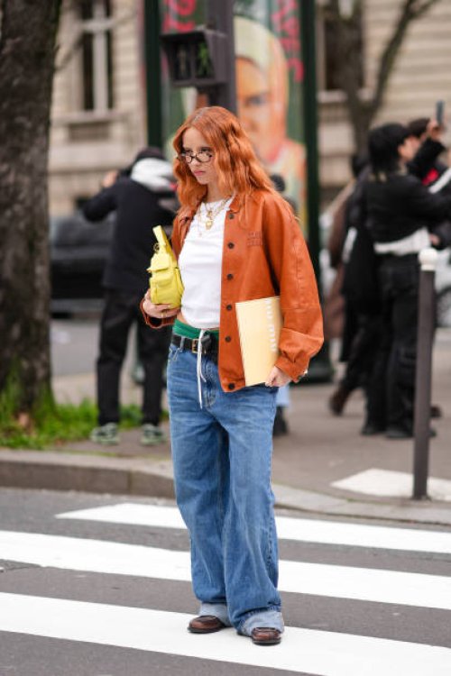Guest wears glasses, an orange leather jacket , a white cropped top, green underwear, a belt, blue wide-leg denim pants / jeans , a yellow leather...