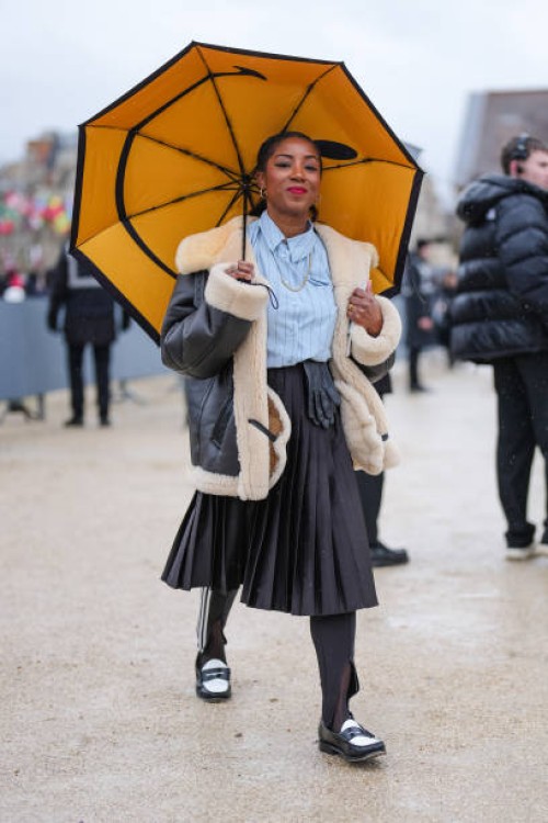 Guest wears gold earrings, gold necklace, light blue lined buttoned up long sleeve shirt, black pleated midi skirt, shiny black leather gloves, shiny...