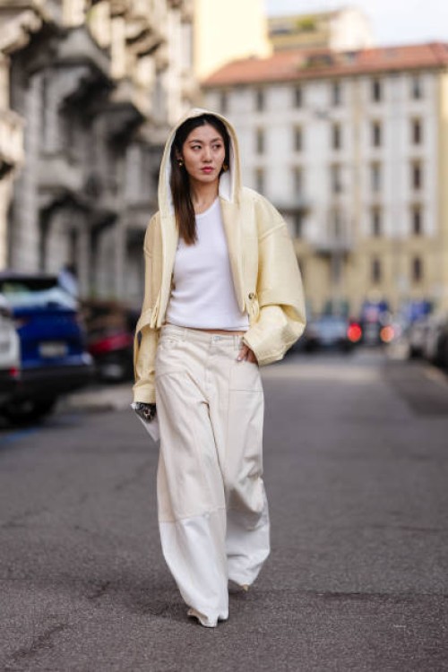 Guest wears gold earrings, light pale yellow hooded cropped leather jacket, white tank top, cream low-rise baggy denim jean pants, outside Sportmax,...