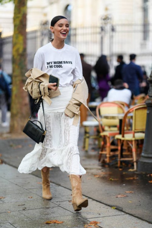 Guest wears gold earrings, white oversized t-shirt with black graphic print, light brown beige cropped trench coat, white sheer mesh lace midi skirt,...