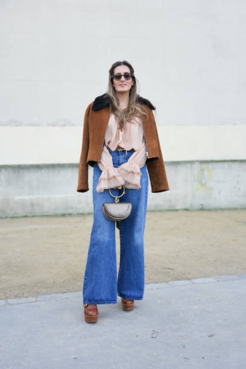 Guest wears gold hoop earrings, dark brown sunglasses, dark brown suede unzipped jacket, light pink long sleeve shirt, navy blue flared denim jean...