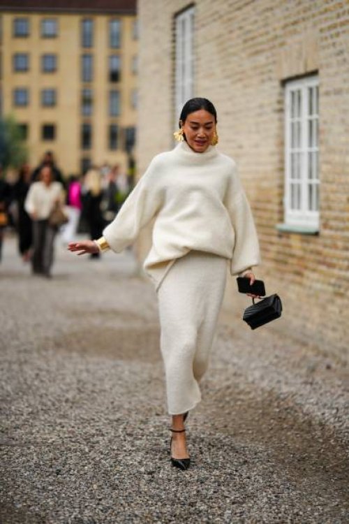 Guest wears gold large earrings, a white latte wool turtleneck pullover, a white latte wool long skirt, a black shiny leather handbag, black shiny...