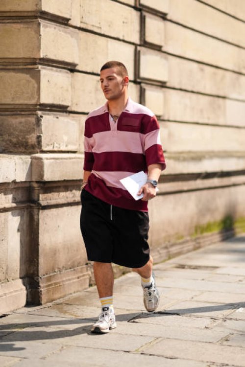Guest wears light pink and dark red striped oversized shirt, a watch, a gold bracelet, a silver bracelet, black shorts, white with yellow striped...