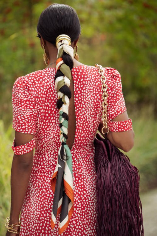 Guest wears long red dress with polkadots, burgundy bag with tassels, golden earrings and braid braided with scarf outside the Marimekko show during...
