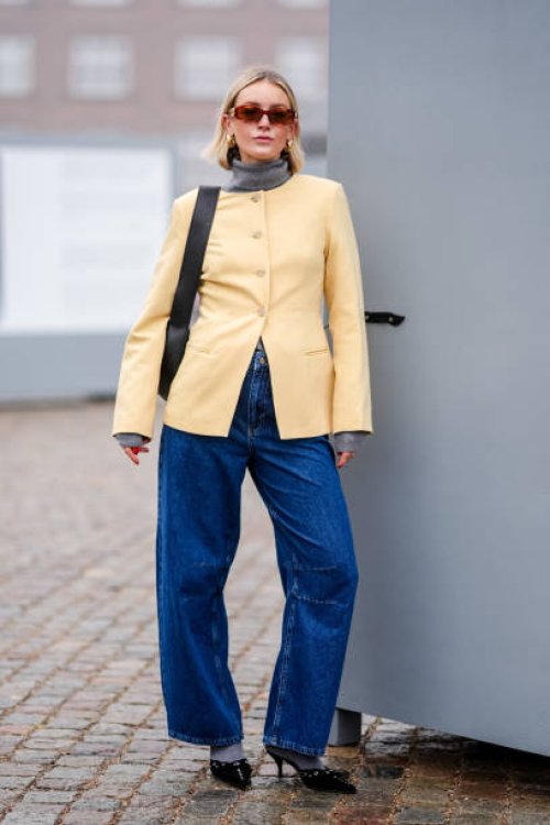 Guest wears Miu Miu sunglasses, golden earrings, a pale pastel yellow jacket, a black leather bag, blue denim jeans pants , pointed shoes, outside...