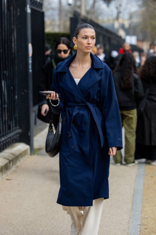 Guest wears navy belted trench coat, black bag, beige pants outside Lacoste during the Womenswear Fall/Winter 2024/2025 as part of Paris Fashion Week...