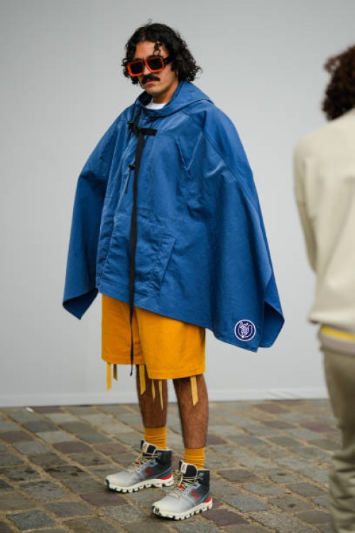 Guest wears orange sunglasses, a blue denim hoodie oversized cloak, yellow orange fringed shorts, yellow socks, white and black gradient print...