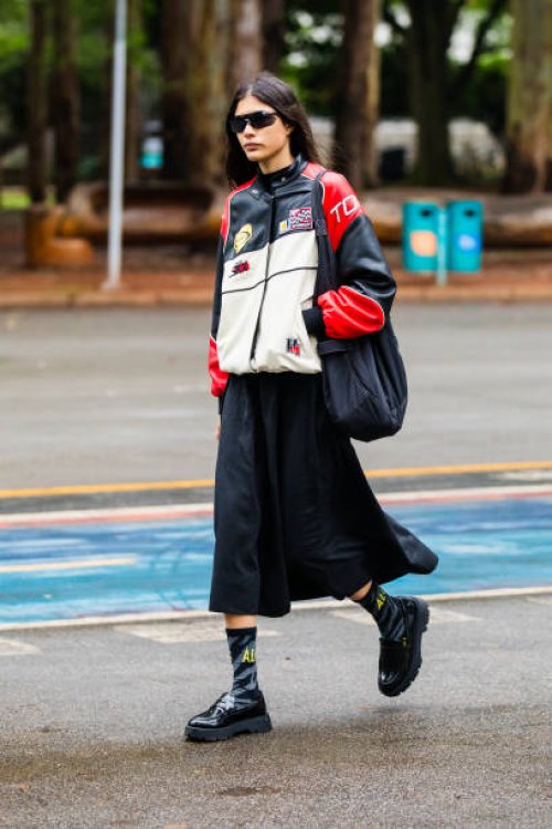 Guest wears racing jacket, black midi skirt, black chunky shoes, black socks with yellow lettering, black oversized bag, black sunglasses at Sao...
