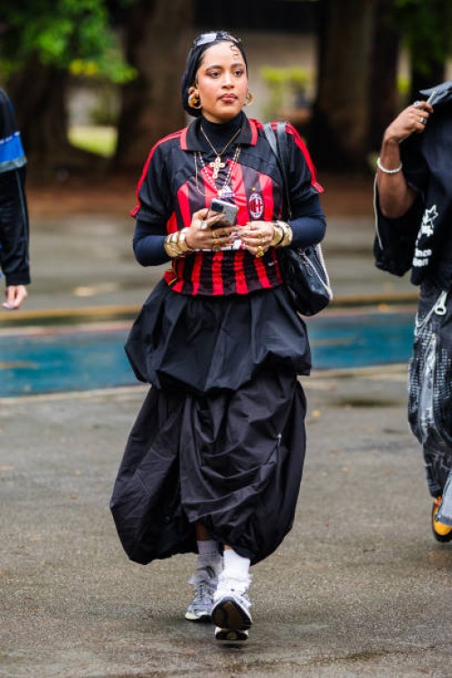 Guest wears red and black football jersey, black pleated skirt, gold jewelry, white socks, chunky sneakers at Sao Paulo Fashion Week 2024 N58 -...