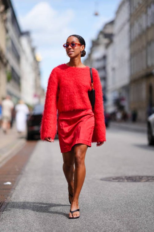 Guest wears red sunglasses, silver hoop earrings, bright red fluffy long sleeve sweater, black leather bag, bright red mini skirt, gold rings, black...