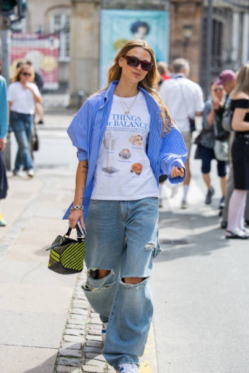 Guest wears ripped denim jeans, blue white button shirt, white tshirt outside Gestuz during day four of the Copenhagen Fashion Week SS25 on August...