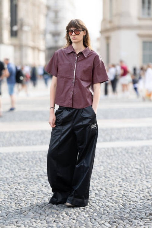 Guest wears satin wide leg trousers, a silver necklace, silver hoop earrings, black sunglasses and a burgundy short sleeved cargo shirt outside...