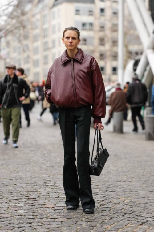 Guest wears shiny burgundy zipped bomber leather jacket, black slim fit flared trouser pants, shiny black Alaïa Le Teckel leather bag, shiny black...