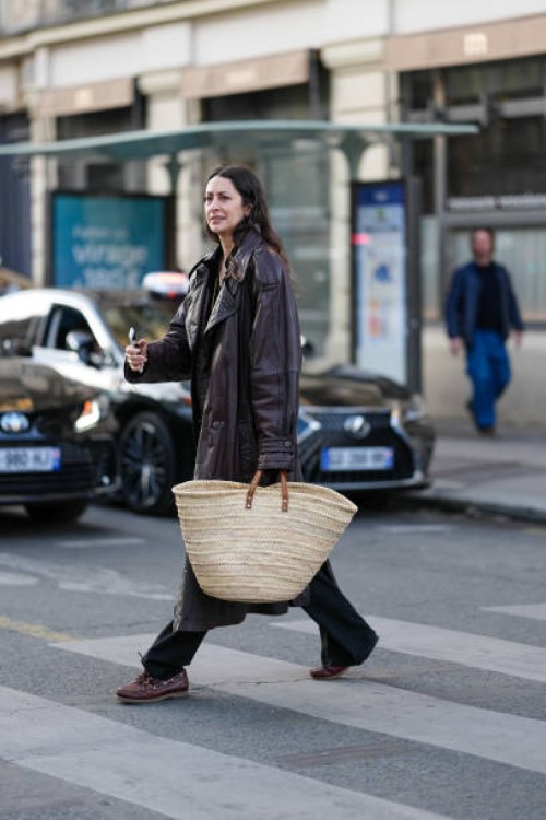 Guest wears shiny dark brown double breasted leather trench coat, light brown basket bag, black relaxed trouser pants, shiny dark brown loafer...