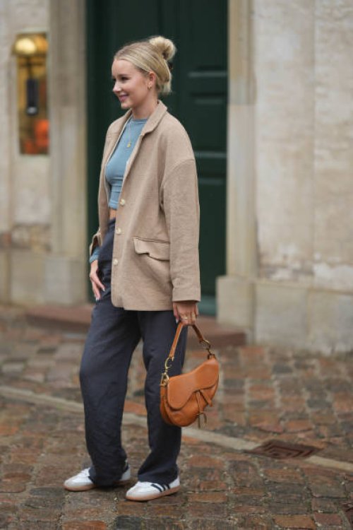 Guest wears silver earrings, a blue gray cropped t-shirt, a beige blazer jacket, gray suit pants, white sneakers, a camel shiny leather Saddle...
