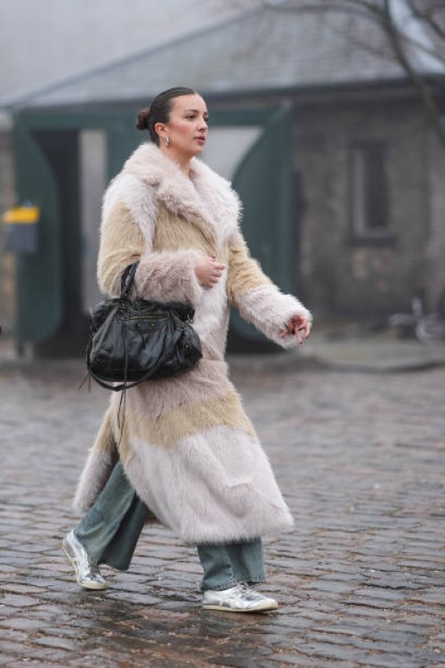 Guest wears silver earrings, a white and beige fluffy faux fur long coat, a black leather Balenciaga bag, flared pants, sneakers, outside Rolf...