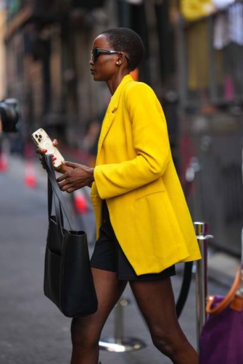 Guest wears sunglasses, a a yellow oversized blazer jacket, a skirt, a black large leather bag, holds an iphone, outside Brandon Maxwell, during New...