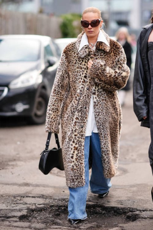 Guest wears sunglasses, a beige leopard print long coat, flared blue denim jeans pants, a black leather bag, outside Skall Studio, during the...