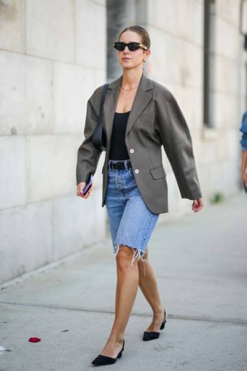 Guest wears sunglasses, a beige oversized blazer jacket, a black low neck top, a belt, blue ripped denim shorts, black pointed flat shoes, outside...