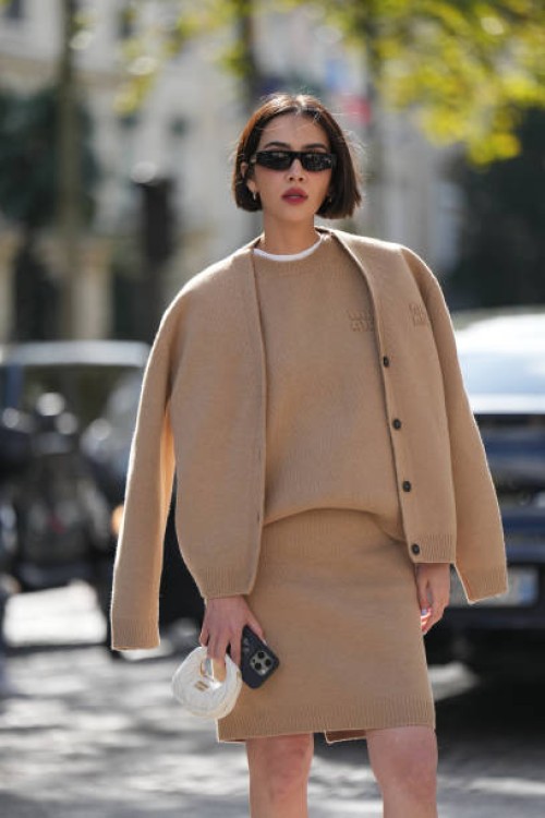 Guest wears sunglasses, a beige pastel / pale brown full look, cardigan, wool pupllover, white t-shirt, mini skirt, a white mini bag, outside Miu...