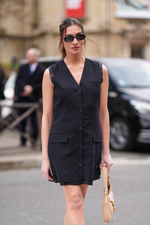 Guest wears sunglasses, a black sleeveless oversized blazer jacket worn as a mini dress , a beige leather bag, outside Miu Miu , during the...