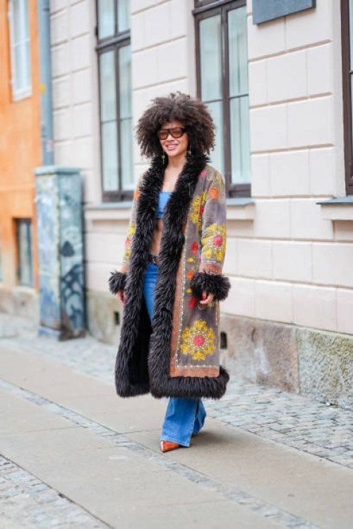 Guest wears sunglasses, a blue bras, a gray and black long winter coat with fluffy parts and printed geometric patterns in yellow and orange, a belt,...