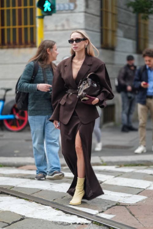 Guest wears sunglasses, a brown oversized blazer jacket as a top, a brown leather belt, a brown leather bag, a long brown skirt with a slit, beige...