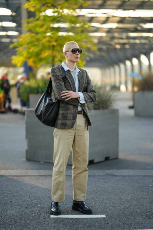 Guest wears sunglasses, a pale blue shirt, a checkered blazer jacket, a black leather bag, a belt, beige pants, black leather shoes, outside COS,...