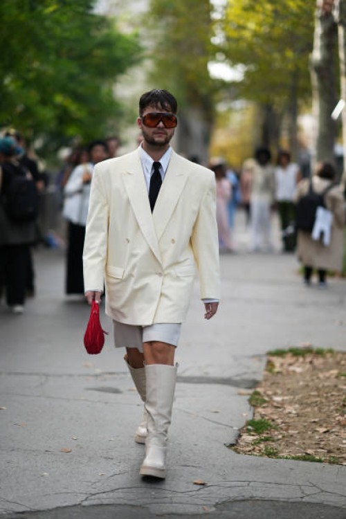 Guest wears sunglasses, a white shirt, a tie, a cream color oversized blazer jacket, white shorts, knee high boots, outside Giambattista Valli,...