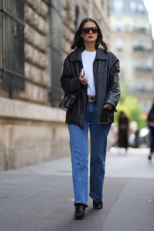 Guest wears sunglasses, a white top, a black leather jacket, a belt, blue jeans, outside Hermes, during the Womenswear Spring/Summer 2024 as part of...
