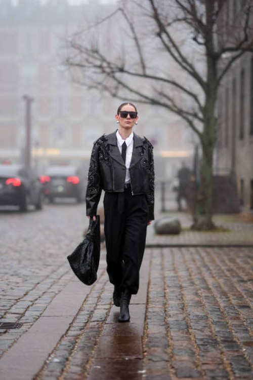 Guest wears sunglasses, earrings, a white shirt, a black tie, a black leather jacket with texture and details, a midi black skirt, a woven leather...