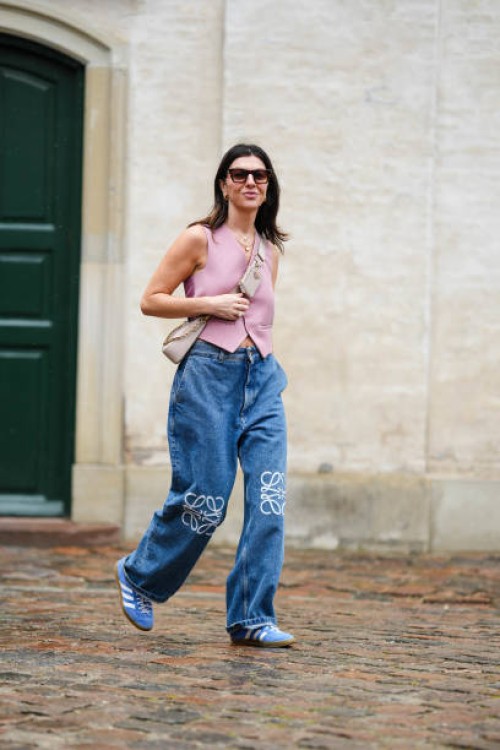Guest wears sunglasses, gold earrings, gold chain necklaces, a pale pink sleeveless buttoned gilet, a beige Epi leather crossbody bag from Prada,...
