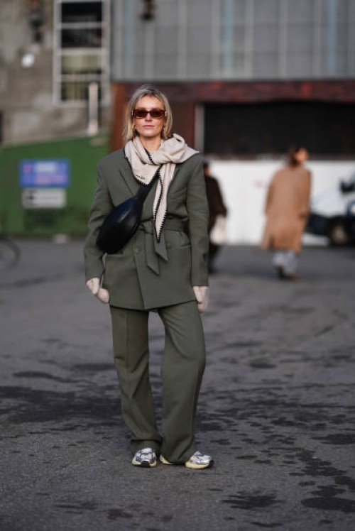 Guest wears sunglasses , golden earrings, a gray scarf, a green khaki oversized blazer jacket , a black leather bag, suit flared pants , sneakers...