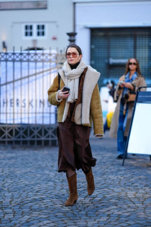 Guest wears sunglasses, golden earrings , a white wool pullover worn as a scarf, a beige aviator jacket with fluffy inner lining , a brown midi...