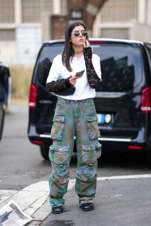 Guest wears sunglasses, necklaces, a white top, black lace mittens, camouflage cargo pants, black leather shoes, outside Anakiki, during the Milan...