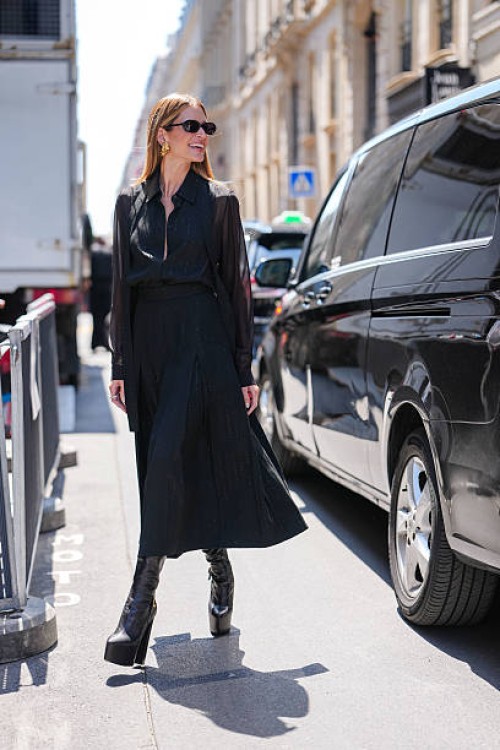 Guest wears sunglasses with dark lenses and gold-toned earrings. The hair is styled straight and loose. A sheer black blouse with long sleeves and a...