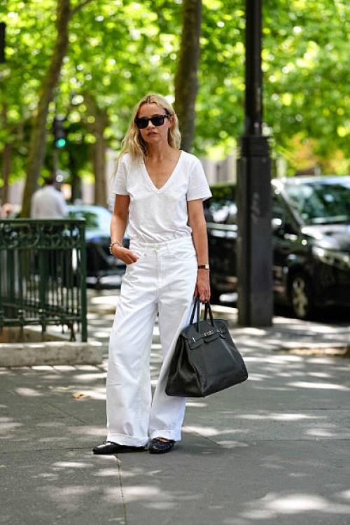 Guest wears sunglasses with dark lenses and a black frame. Hair is styled in loose waves. A white V-neck T-shirt is paired with high-waisted white...