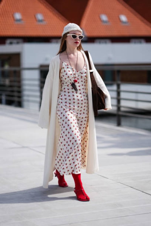 Guest wears white knit hat, cream sunglasses, black necklace red pendant, cream midi dress with red floral detail, cream cardigan trench sweater,...