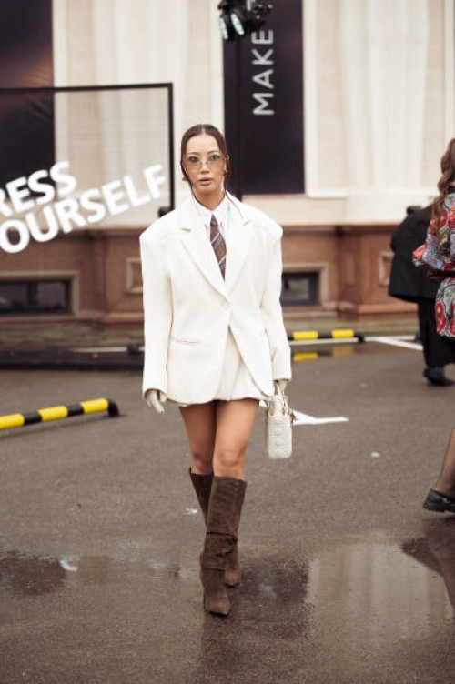 Guest wears white mini skirt, white shirt, brown stripped tie, white blazer, white Dior bag and long brown boots during the 10th season of the Visa...