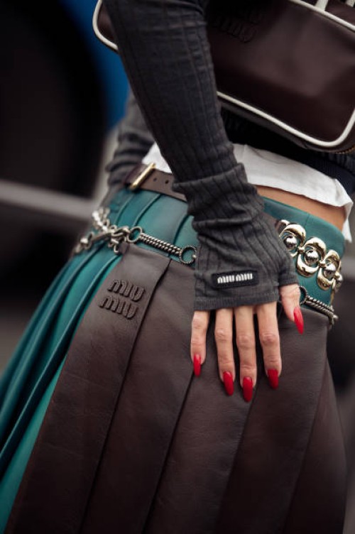 Guest wears white shirt, black leather jacket, brown and green pleated leather midi skirt with chain belts, brown Miu Miu bag outside the Miu Miu...