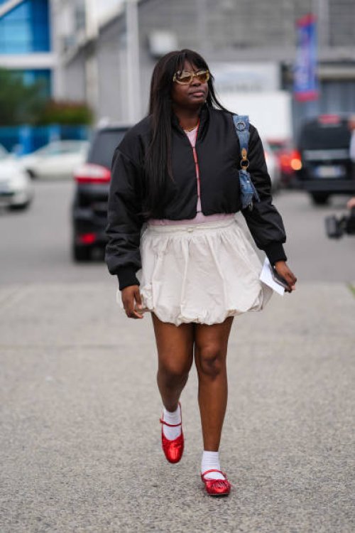 Guest, wears yellow lined glasses with dark brown lens, black cropped bomber jacket, pink shirt, white skirt, white socks, red ballet shoes,outside...