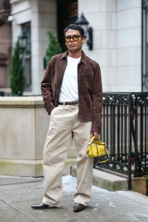Guest wears yellow sunglasses, a brown suede jacket , a white t-shirt, a belt, beige pants, brown leather pointed shoes, a yellow Loewe bag, outside...