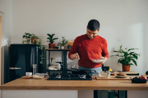 handsome cheerful man standing in a kitchen preparing a meal - food stock pictures, royalty-free photos & images