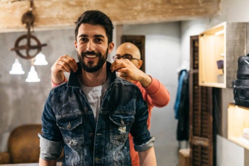 handsome man trying on new denim jacket with stylist in modern boutique - fashion stock pictures, royalty-free photos & images