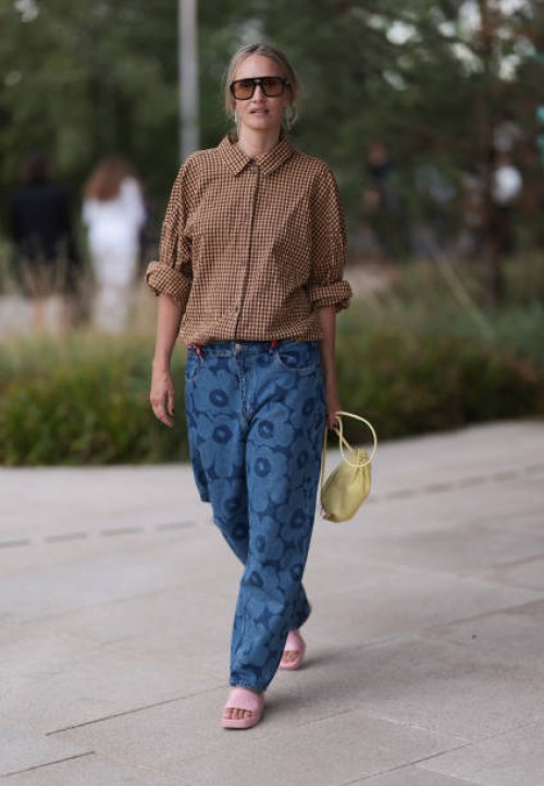 Hanni Gohr is seen wearing angular aviator sunglasses with a black frame and orange lenses, white dangling chain earrings, a buttoned brown and beige...