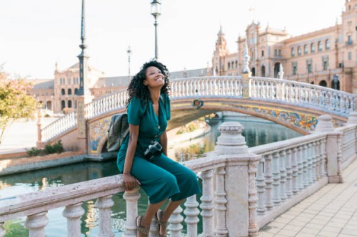 happy afro woman sitting on railing at plaza de espana, seville, spain - travel stock-fotos und bilder