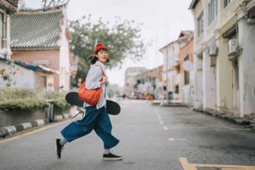 happy asian chinese young woman crossing road carrying skateboard in old town - travel stock pictures, royalty-free photos & images