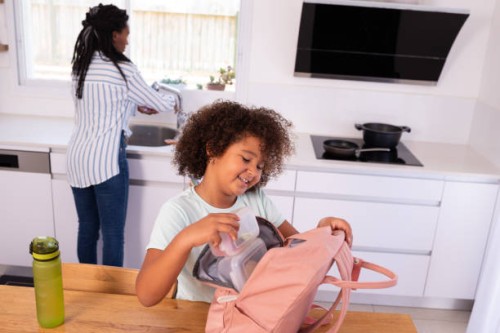 happy elementary-age girl putting a lunch box into a pink backpack before going to school. - junk food stock pictures, royalty-free photos & images