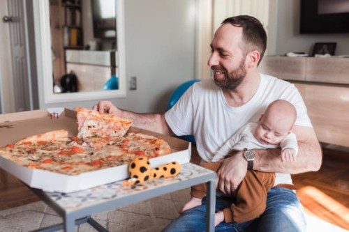 happy man eating pizza while the baby sleeps in his arms - junk food stock pictures, royalty-free photos & images