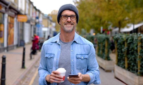 happy man outdoors using his cell phone while drinking a cup of coffee - junk food stock pictures, royalty-free photos & images